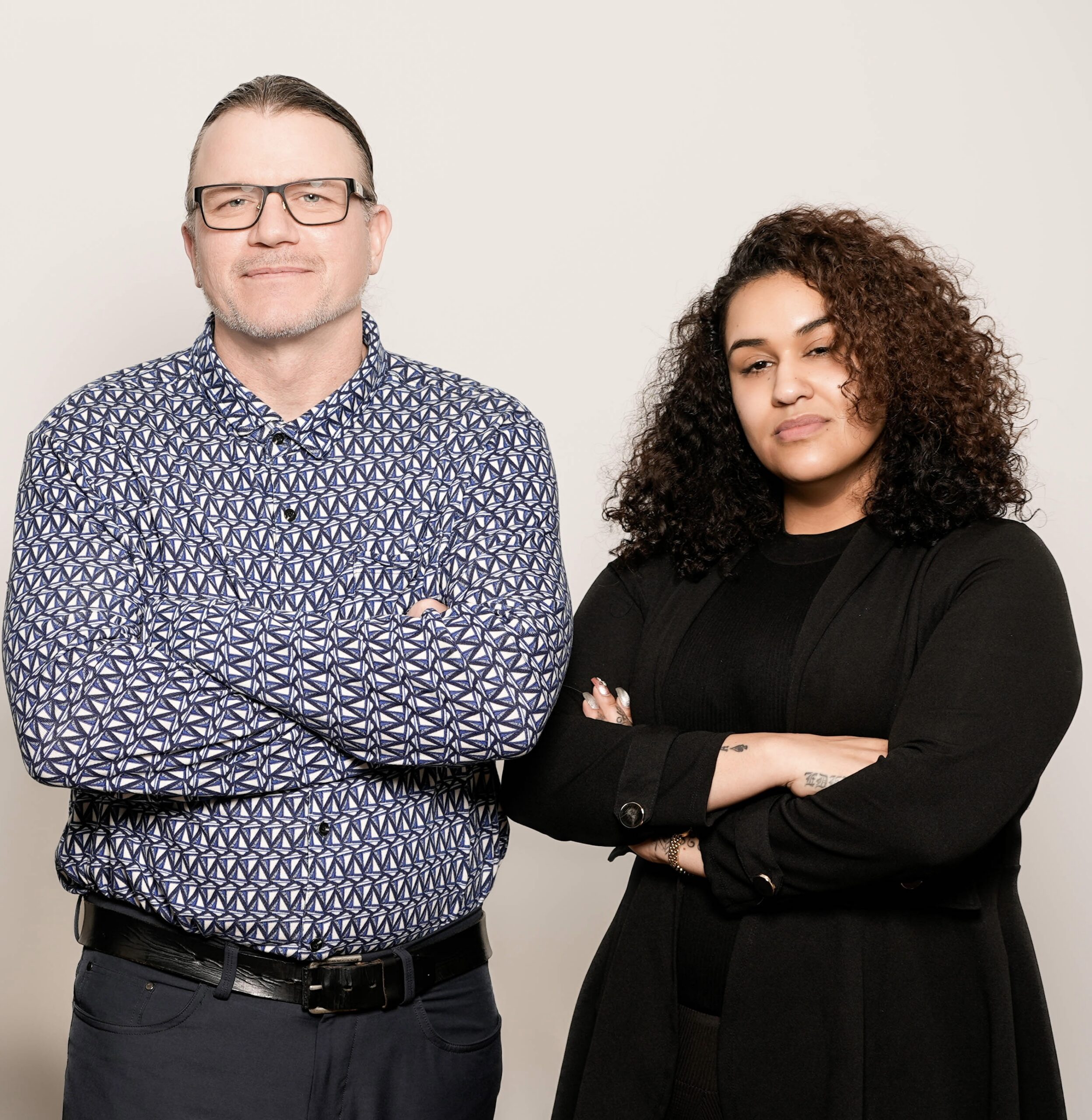 Man in patterned blue shirt and glasses standing with woman in black outfit with curly hair, both with arms crossed